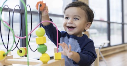 An adorable toddler boy sits at a table in a doctor's waiting room and reaches up cheerfully to play with a toy bead maze.